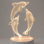 A 3D optical illusion LED night light featuring two dancing dolphins in a wireframe design, glowing with a warm yellow light on a white cylindrical base against a neutral gray background.