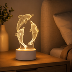 A 3D optical illusion LED night light depicting two dancing dolphins, shown illuminated on a wooden bedside table in a cozy bedroom setting.