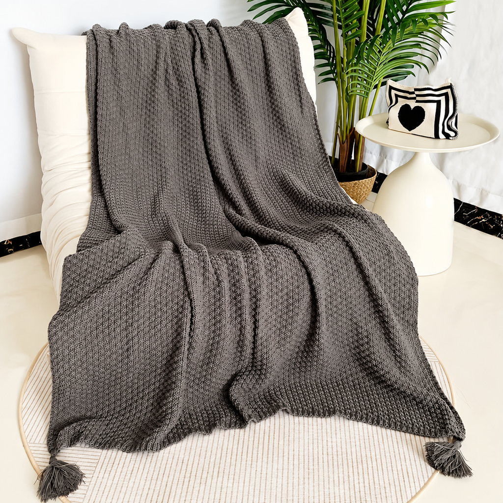 Gray acrylic knit throw blanket spread over a cream chair with corner tassels visible