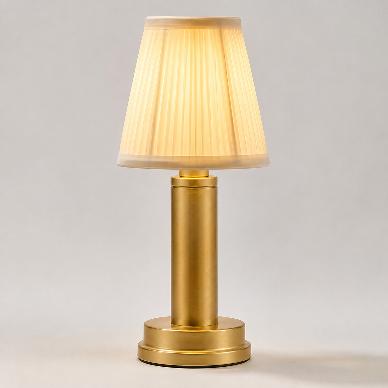 Matte Gold table lamp with a pleated cream shade glowing against a plain light-gray studio background.