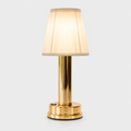 Gold table lamp with a pleated cream shade glowing against a plain light-gray studio background.