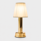 Gold table lamp with a pleated cream shade glowing against a plain light-gray studio background.