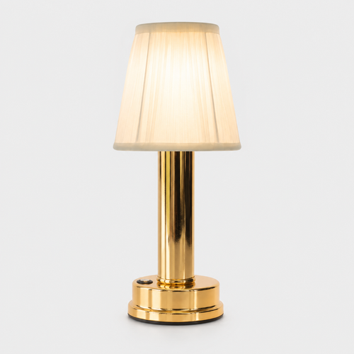 Gold table lamp with a pleated cream shade glowing against a plain light-gray studio background.