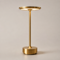 Gold table lamp with a round shade, slim stem, circular base, and warm light on a plain background.