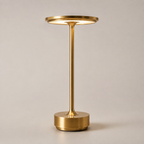 Gold table lamp with a round shade, slim stem, circular base, and warm light on a plain background.