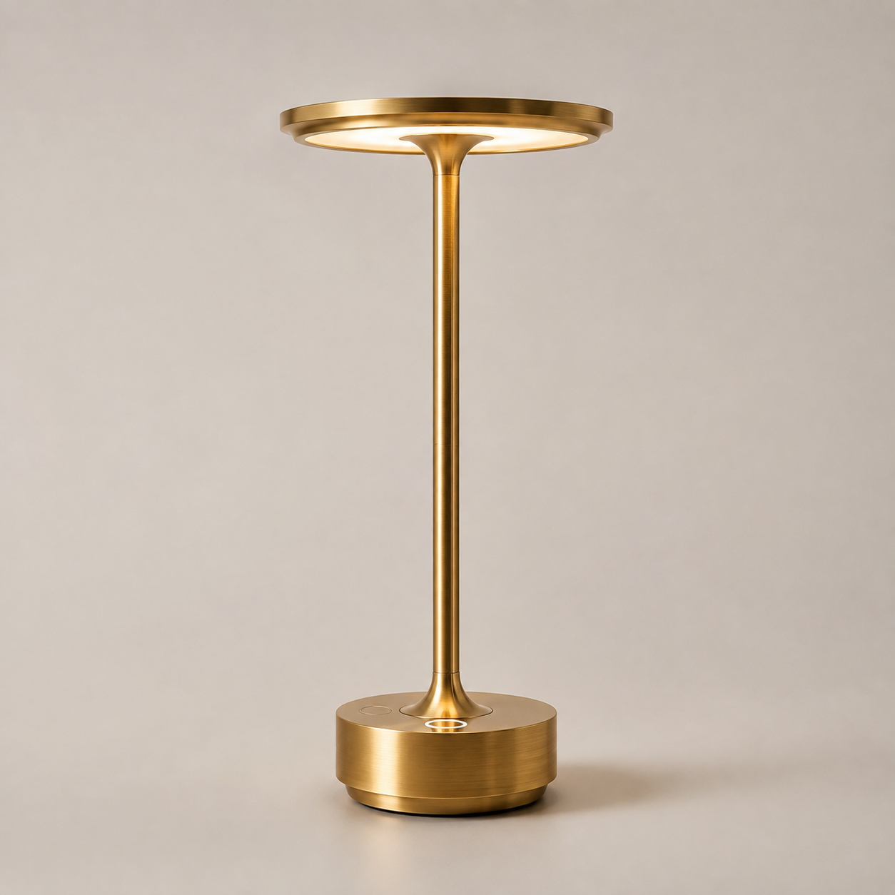 Gold table lamp with a round shade, slim stem, circular base, and warm light on a plain background.