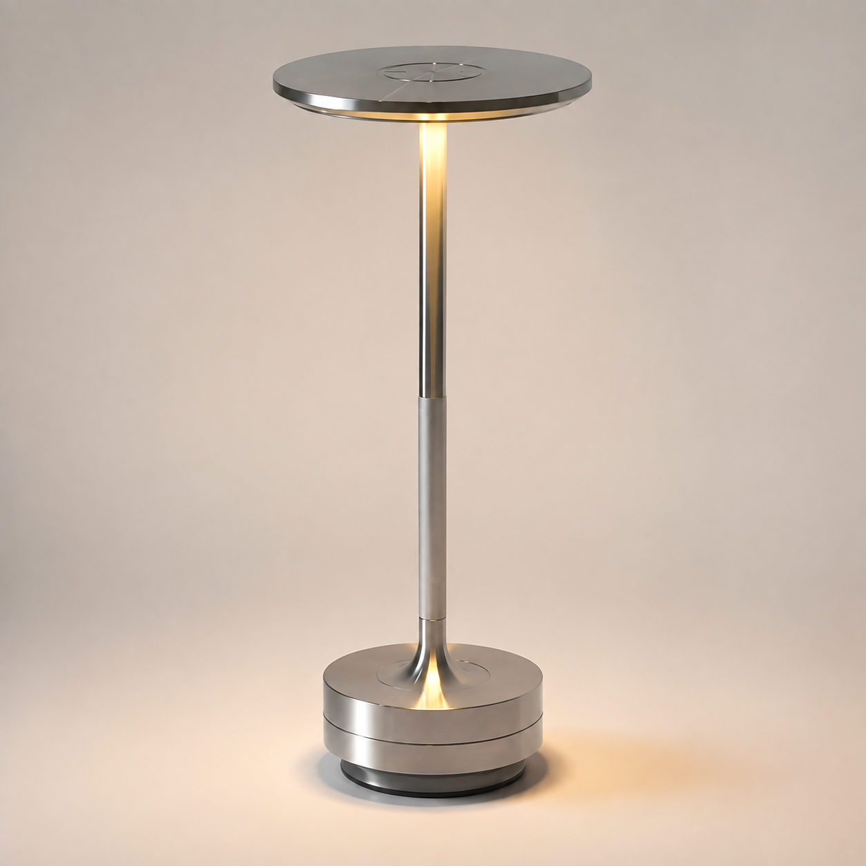 Silver table lamp with a round shade, slim stem, circular base, and warm light on a plain background.
