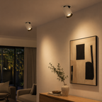 Two modern white cylindrical spotlight ceiling lights mounted on a beige wall in a contemporary living room setting, casting a warm glow downward.
