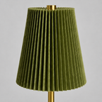 An olive green pleated fabric lampshade with a gold metal finial on top and a matching gold stem, shown against a neutral gray background.