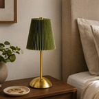 A table lamp with a pleated olive green shade and a polished gold brass base, styled on a dark wood nightstand next to a bed in a cozy bedroom setting.