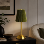 A modern table lamp with a pleated olive green fabric shade and a brushed gold metal base, styled on a dark wooden side table in a cozy living room setting.