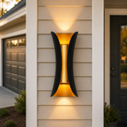 Black and gold outdoor wall sconce glowing on beige siding between a garage and front door.