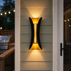 Black and gold outdoor wall sconce casting warm beams on gray siding beside a deck at dusk.