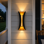 Black and gold outdoor wall sconce glowing on gray siding beside a dark doorway at dusk.