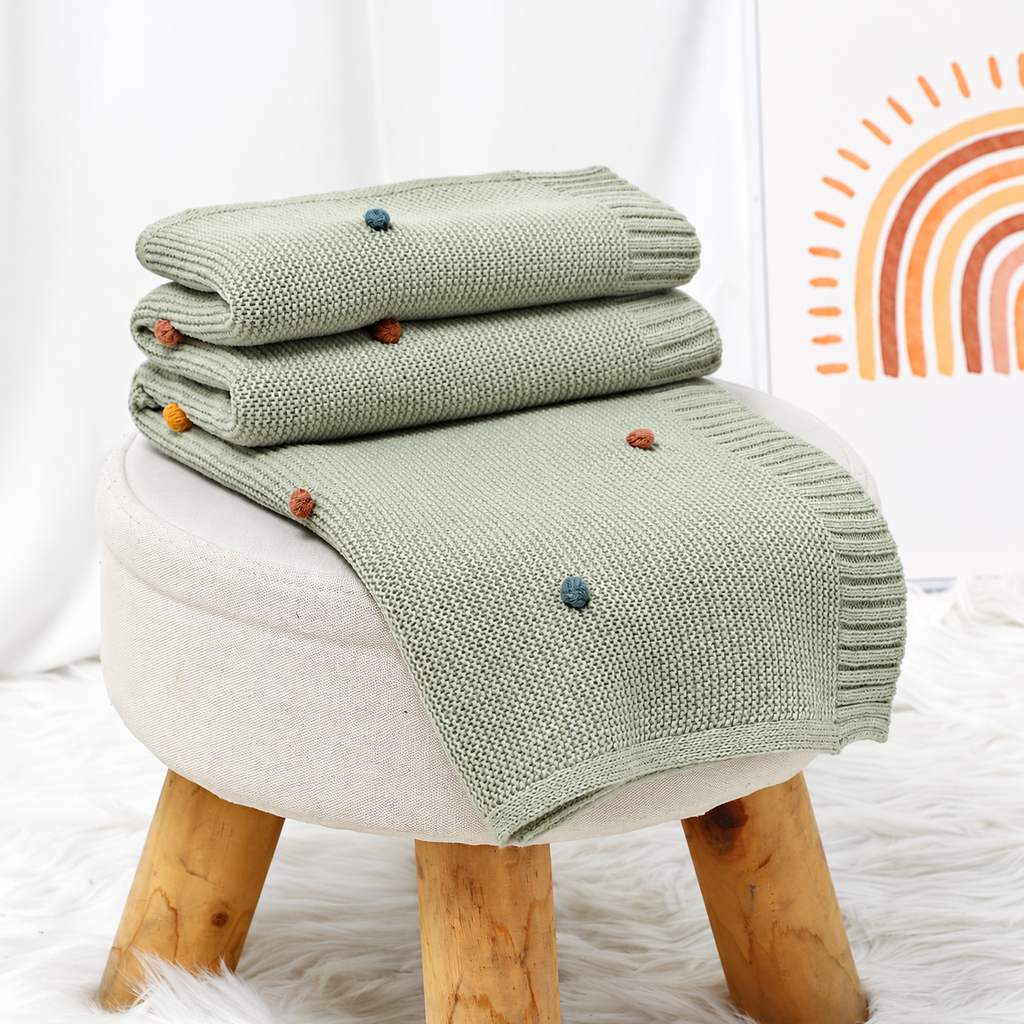 Light green acrylic knit throw blanket with colorful pom pom dots arranged on a stool