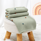 Light green acrylic knit throw blanket with colorful pom pom dots arranged on a stool