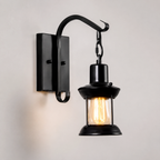 Black lantern wall sconce with a curved arm and exposed warm glowing bulb against a neutral wall.
