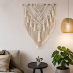Natural macrame wall hanging above a small black table and green plants.
