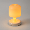 A modern mushroom-shaped table lamp with a glowing warm yellow-orange shade and a matte cream-colored base, set against a neutral gray background.
