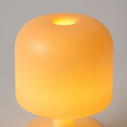 A modern, rounded cylindrical table lamp glowing with a warm yellow-orange light against a neutral gray background.