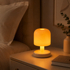 A small mushroom-shaped table lamp glowing with warm yellow light on a bedside table next to a bed, with a vase of greenery and a book in the background.