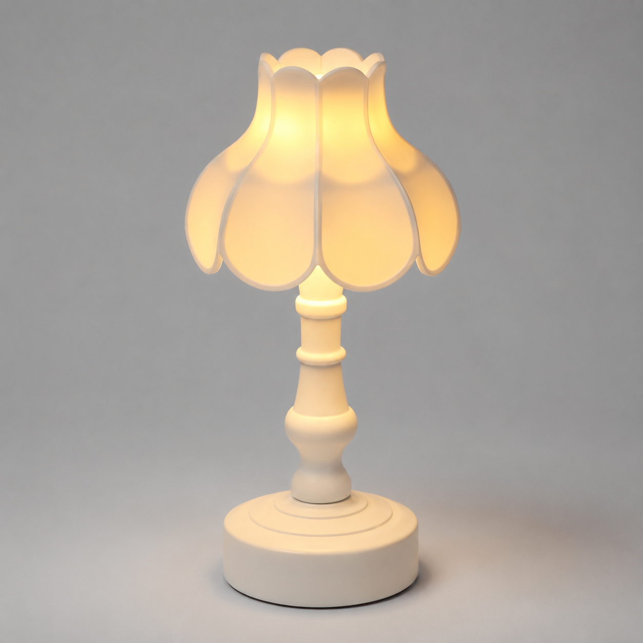 A small table lamp with a warm white ceramic base and a scalloped petal-shaped shade that glows with a soft yellow light, set against a neutral gray background.