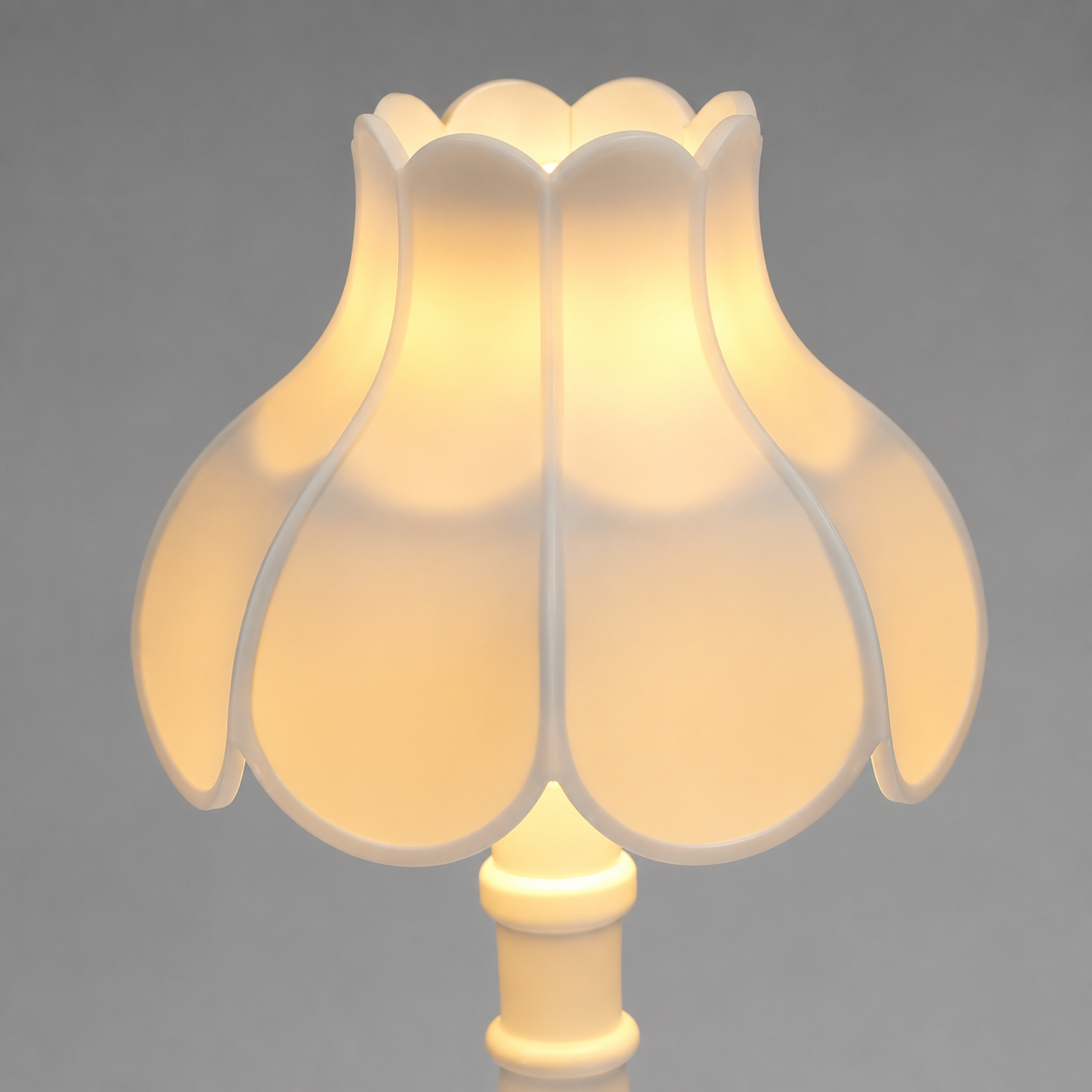 A close-up of a warm white or cream-colored tulip-shaped lampshade on a matching lamp base, glowing with a soft warm yellow light against a neutral gray background.
