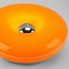 A close-up of a glossy orange circular product, possibly a lamp or decorative object, featuring a metallic silver dome in the center, set against a neutral gray background.