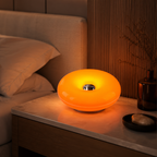 A glowing orange circular tabletop lamp sits on a bedside table next to a bed in a warmly lit bedroom.