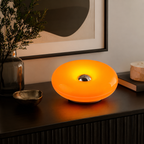 A modern circular LED night light with a glowing orange light and a metallic silver center, styled on a dark wooden console table in a home interior setting.