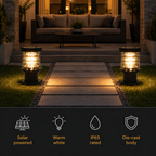 Two solar-powered outdoor bollard lights glowing with warm white light on either side of a stone path in a backyard setting. A dark informational banner at the bottom features icons and text for 'Solar powered', 'Warm white', 'IP65 rated', and 'Die-cast body'.