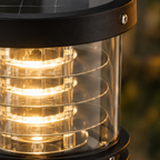 Close-up of a solar-powered outdoor lantern with a black top cap, clear cylindrical body, and warm yellow light glowing through internal rings.