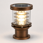 A solar-powered outdoor bollard light with an antique bronze metal finish, clear glass shade, and integrated solar panel on top, shown illuminated against a neutral light gray background.