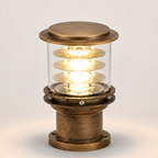 A vintage-style industrial table lamp with a brass-colored metal base and top cap, a clear glass cylinder, and a glowing spiral filament bulb inside, set against a plain light gray background.