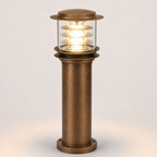 A modern industrial-style column light fixture with a brushed bronze metal body, clear glass shade, and glowing warm white LED light, shown against a neutral light gray background.