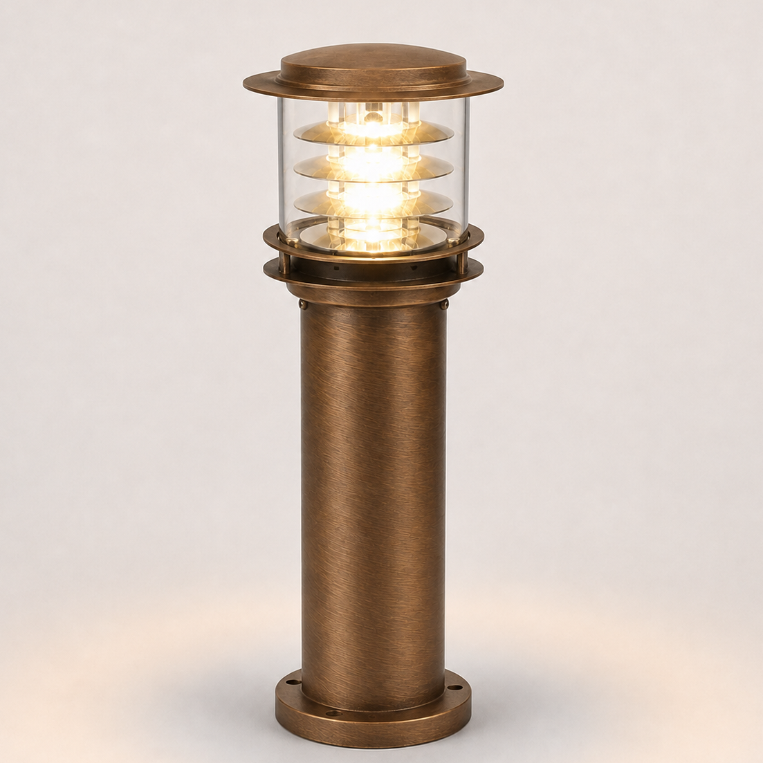 A modern industrial-style column light fixture with a brushed bronze metal body, clear glass shade, and glowing warm white LED light, shown against a neutral light gray background.