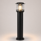 A modern black outdoor bollard light with a clear glass shade and warm glowing LED light, set against a neutral light gray background.