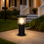 A black solar-powered outdoor bollard light with a clear glass shade, glowing with warm light on a concrete walkway in front of a house.