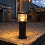 A cylindrical outdoor bollard light with a black metal body and glowing warm yellow glass, placed on a wooden deck at dusk near a patio grill and dining table.