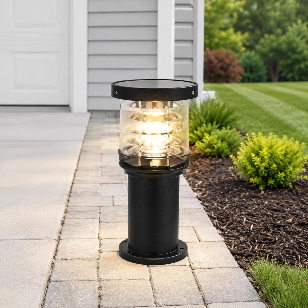 A black outdoor bollard light with a clear glass shade and warm glowing light inside, positioned on a stone pathway in a landscaped yard next to a house.