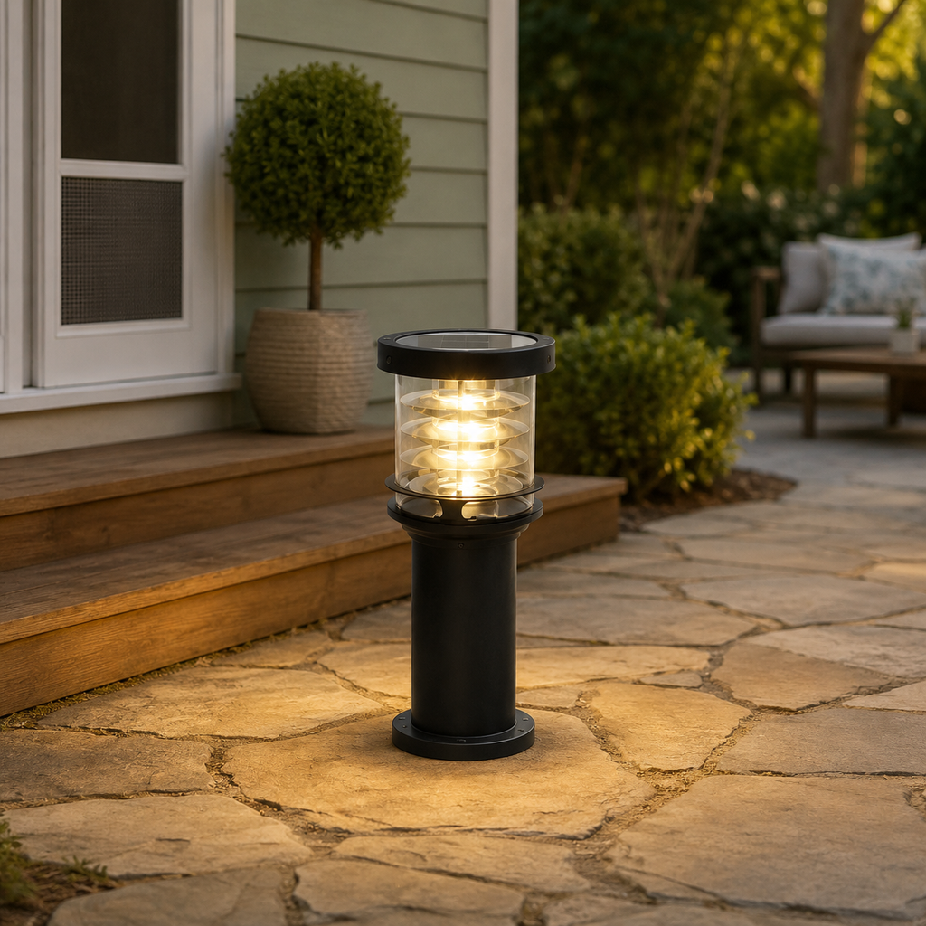A black outdoor solar post light with a clear glass shade is turned on, casting a warm yellow glow onto a stone patio in front of a house.