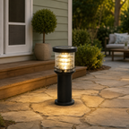 A black outdoor solar post light with a clear glass shade is turned on, casting a warm yellow glow onto a stone patio in front of a house.