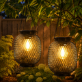 Two black outdoor solar lanterns glow above a leafy backyard at dusk.
