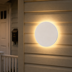 White round outdoor wall light glowing on horizontal gray siding beside a doorway.