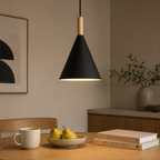 Black conical pendant light with a natural wood stem, glowing warm white above a wooden dining table with pears, a mug, and books.
