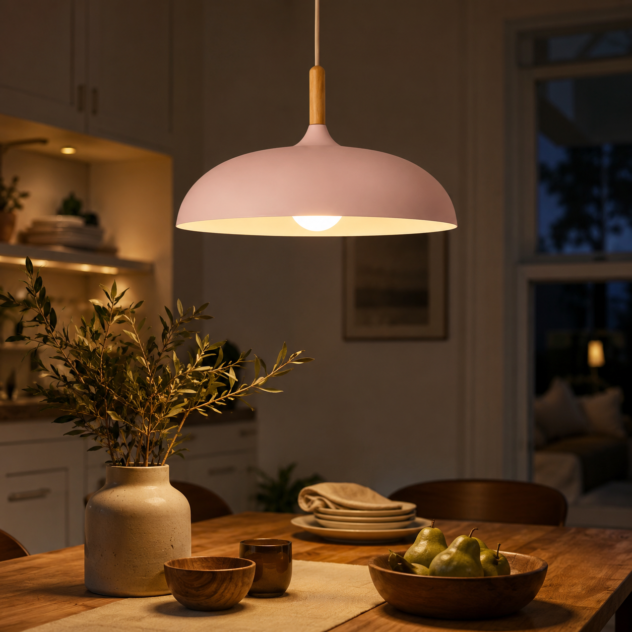 Pink wide-dome pendant light with a natural wood stem, glowing warm white above a wooden dining table with fruit, tableware, and a vase.
