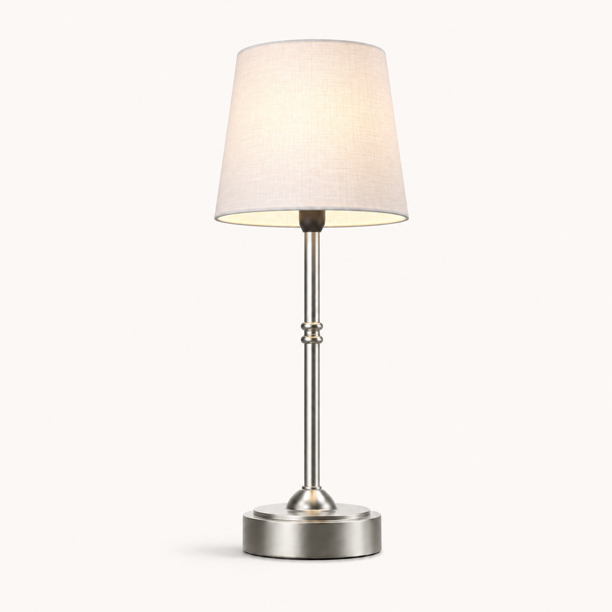 A modern table lamp with a tapered off-white fabric shade, a brushed silver metal stem, and a round silver base, shown lit against a plain off-white background.