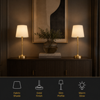 Two matching table lamps with cream fabric shades and gold bases are shown on a dark wood cabinet in a styled interior setting. A framed abstract print and a vase with greenery hang above, and a black banner at the bottom lists product features: fabric shade, gold finish, slim profile, and warm glow.