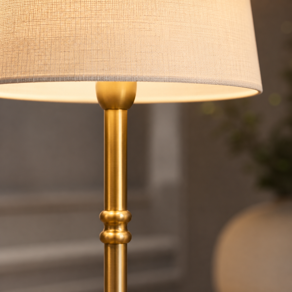 A close-up of a floor lamp with a warm beige linen-textured drum shade and a polished gold brass stand, shown lit against a softly blurred indoor background.