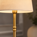 A close-up of a floor lamp with a warm beige linen-textured drum shade and a polished gold brass stand, shown lit against a softly blurred indoor background.